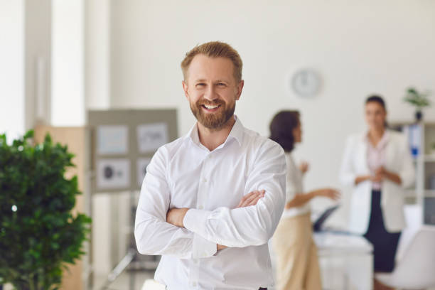 Happy successful businessman or company employee standing in office looking at camera Portrait of happy successful handsome businessman in office workspace. Confident bearded businessman or head of company department standing arms crossed looking at camera and smiling founder photos stock pictures, royalty-free photos & images