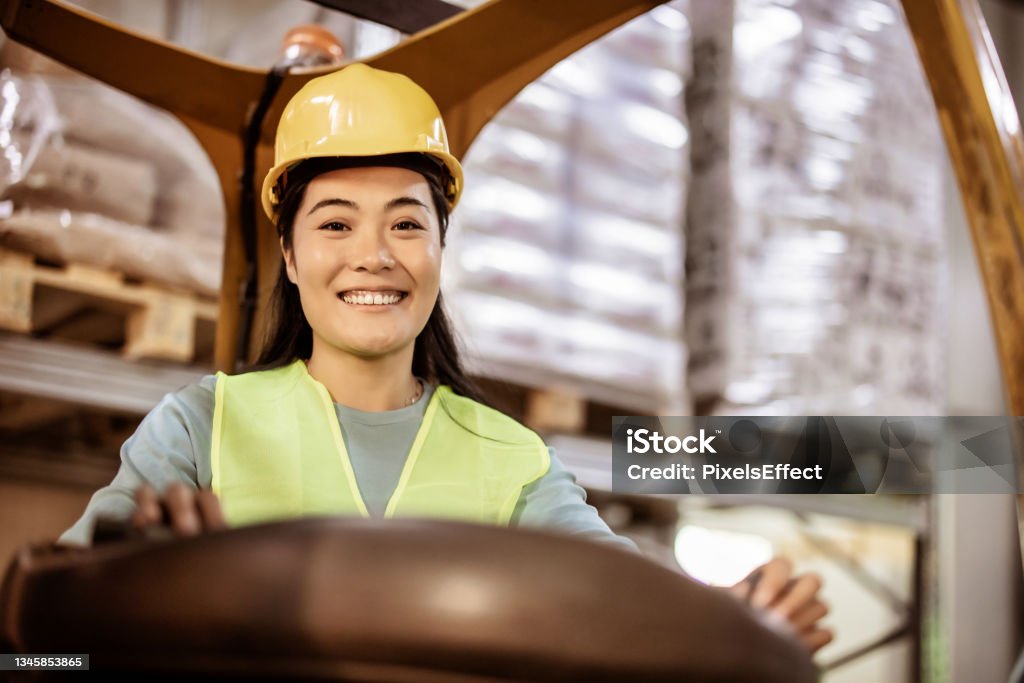 Let’s get things moving Beautiful Asian female worker looking at camera while sitting in forklift in warehouse. This is a freight transportation and distribution warehouse. Industrial and industrial workers concept Packaging Stock Photo