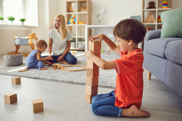 lindo niño pequeño construyendo una torre de bloques de madera con su hermano pequeño y mamá de fondo - cube baby child block fotografías e imágenes de stock