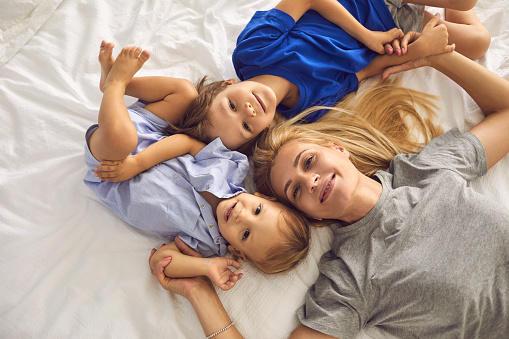 Portrait of a beautiful smiling young blonde mother with two cute little sons lying on the bed and looking at the camera. Top view. Concept of happy motherhood.