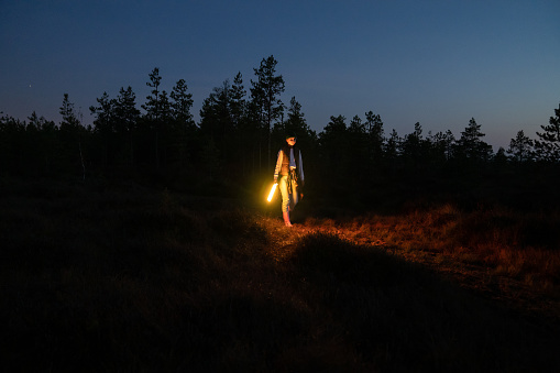 Young woman walk in nature at night holding led lamp to highlight pathway from forest to camp, village or car after hiking. Casual female in rubber boots wondering autumn field or swamp after sunset