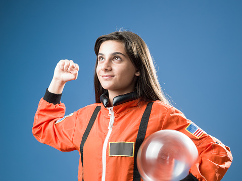 Portrait of teenager girl wearing orange astronaut costume standing in front of blue colored wall. She has long brown hair and holding a glass helmet. Shot in studio with a full frame mirrorless camera.