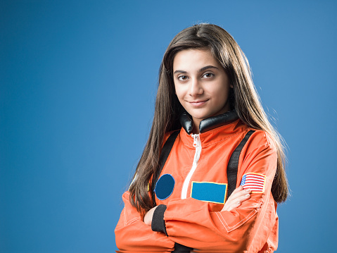 Portrait of teenager girl wearing orange astronaut costume standing in front of blue colored wall. She has long brown hair. Shot in studio with a full frame mirrorless camera.
