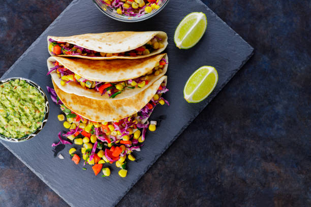 istock Vegetarian tacos with various vegetables, guacamole and sliced lime on dark background. Tacos with sweet corn, purple cabbage and pepper on a slate board. Copy space 1345872700