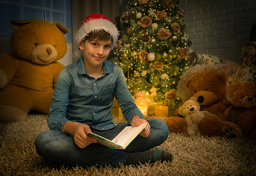 Happy boy with santa's hat reading book sitting on the floor in front of the Christmas tree and Teddy bears