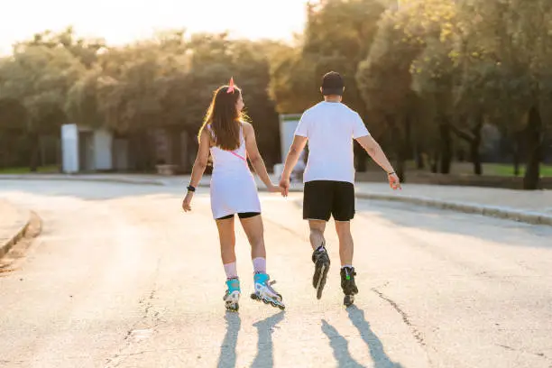 Couple in their backs skating on inline skates holding hands on a street in the middle of a park