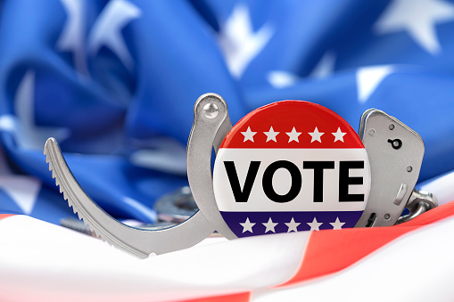 Images depicts a voting button against handcuffs and the American flag, as voter fraud is more prevalent than ever and those caught will be arrested.