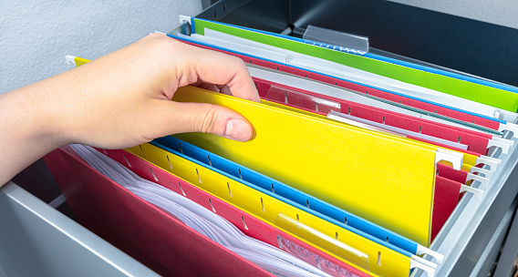 Person's hand picks yellow hanging file folder from open drawer. Administration clerk finding paperwork from office storage. Corporate information record, organized category and database management concept.