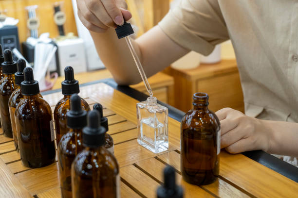 Osoba ostrożnie przelewająca płyn do butelki perfum otoczonej innymi butelkami na drewnianym stole.