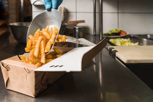 Hand of an unrecognizable cook serving french fries in box for customer in restaurant.