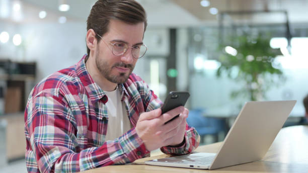 istock Professional Young Man Browsing Internet on Smartphone and Laptop in Cafe 1345943450