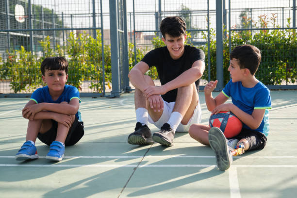 un allenatore siede sul pavimento del campo da calcio riposando e parlando con due studenti. - soccer child indoors little boys foto e immagini stock