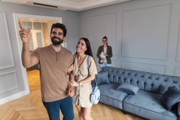 Happy couple enjoying in real estate showing. Young happy couple exploring the apartment during real estate showing while man is aiming at something. Real estate agent is in the background. Copy space. looking around stock pictures, royalty-free photos & images