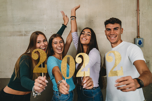 Happy new year 2022 with friends. Four friends are celebrating together the coming of the new year. They are smiling, looking at camera. They are holding a glitter 2022 number.