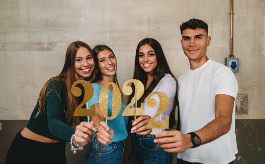Happy new year 2022 with friends. Four friends are celebrating together the coming of the new year. They are smiling, looking at camera. They are holding a glitter 2022 number.