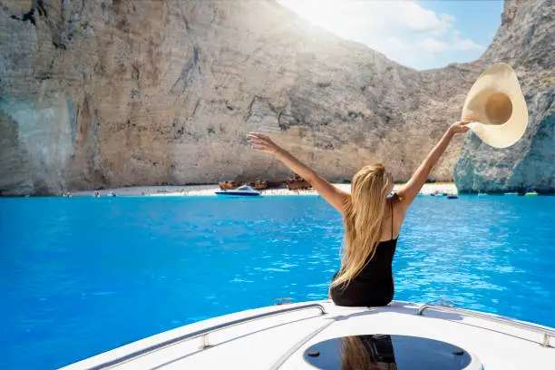A happy tourist woman sitting on a boat enjoys the beautiful view to the turquoise sea of Shipwreck Navagio Beach, Zakynthos island, Greece
