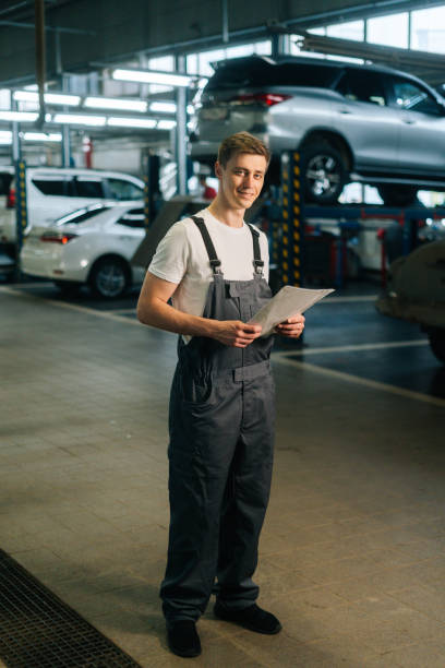 Vertical portrait of smiling handsome young mechanic male wearing uniform holding clipboard standing in auto repair shop garage. Vertical portrait of smiling handsome young mechanic male wearing uniform holding clipboard standing in auto repair shop garage, with vehicle background, looking at camera car portrait men expertise stock pictures, royalty-free photos & images