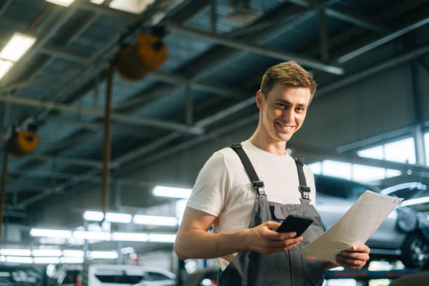 seitenansicht des lächelnden, gutaussehenden jungen mechanikers, der eine uniform trägt, die zwischenablage hält und ein sms-mobiltelefon verwendet - mechanic auto repair shop manual worker men stock-fotos und bilder