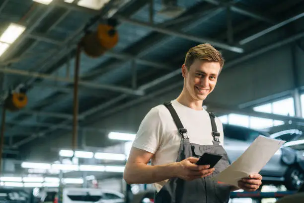 Side view of smiling handsome young mechanic male wearing uniform holding clipboard and using texting mobile phone, standing in auto repair shop garage, with vehicle background, looking at camera