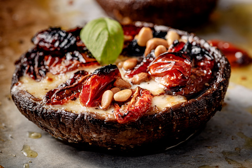 Close-up view of a large Portobello mushroom, filled with sun-dried tomatoes, mozzarella and pine nuts topped with fresh basil. The mushrooms have their stalks removed and the tops and bottoms are brushed with olive oil. The sun dried tomato and mozzarella mix is spooned into the mushroom and topped with some pine nuts and baked for about 20 mins, the finished off with some fresh basil leaves. A delicious and easy vegetarian lunch or light dinner, colour, horizontal format with some copy space