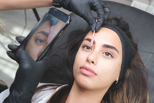 Beautician is using a eyebrow brush and taking a photo with smart phone. Beautiful young woman in beauty salon on eyebrow makeup treatment. Beautician doing eyebrows tattooing. Ruler
