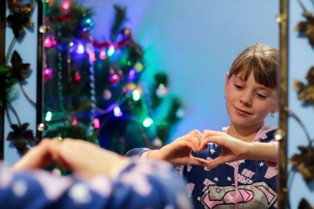 defocus little blonde caucasian girl 10 years old in pajamas shows gesture heart on mirror reflection. portrait on mirror. reflection. new year. childhood. out of focus - 10 11 years child human face female imagens e fotografias de stock