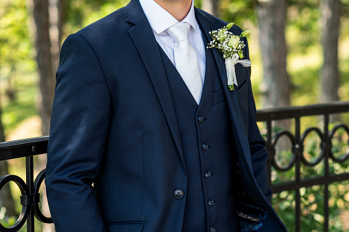Unrecognizable young groom in elegant dark blue suit with beautiful white roses boutonniere. Groom fashion detail shot.