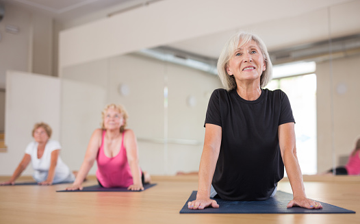 Smiling sporty mature woman with group of females exercising Hatha yoga poses in modern yoga studio, doing Upward Facing Dog stretching pose