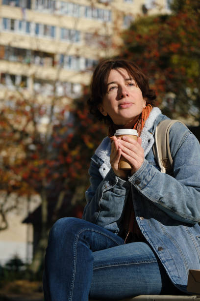 istock Mature woman is drinking coffee while waiting for someone on park bench 1345997835