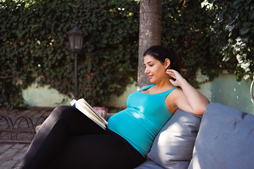 A Young pregnant woman sits in the backyard and reads a book.
