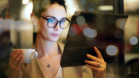 Stock image of a professionally dressed city woman wearing glasses. She’s in a coffee bar in the evening, checking messages on her digital tablet.