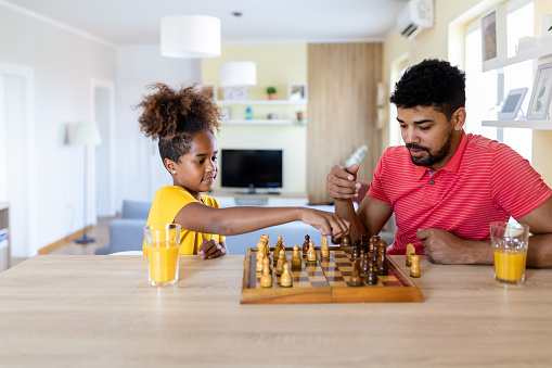 Cute african american girl playing chess with father at home. Little daughter thinking over next move, playing chess with father, strategy