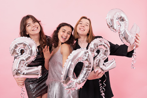 A company of cheerful girlfriends on a pink studio background with silver balloons in the form of the numbers 2022.
