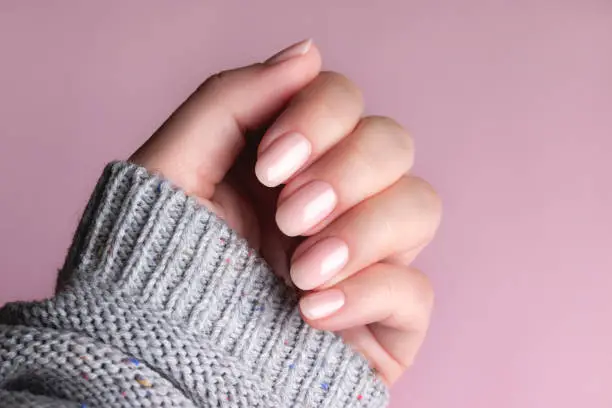 Female hand in gray knitted sweater with beautiful manicure - pink nude nails on pink background. Nail care concept