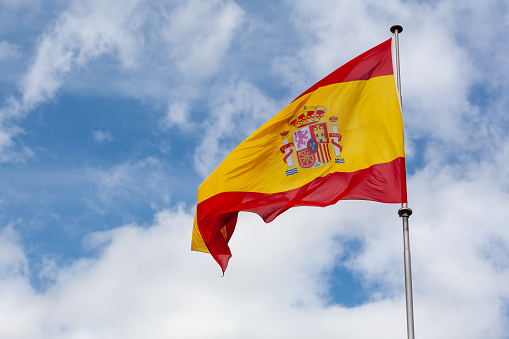 A large flag of Spain with the colors red and yellow, the national insignia, flutters in the wind against the background of the blue sky with clouds.