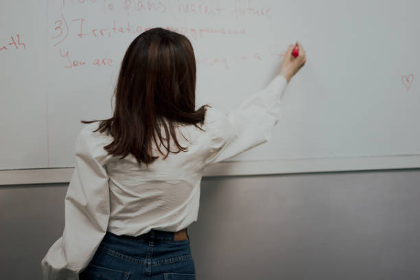 istock Back view of a cute young englisg language teacher in white blouse writing on a board and giving a class 1346021019