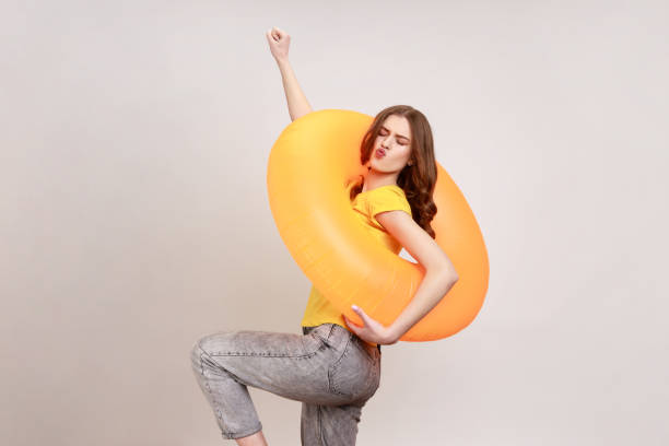 портрет беззаботной игривой привлекательной женщины молодого возраста в желтой футболке и джинсах, поднимающих руку, позирует с резиновым - ring buoy стоковые фото и изображения