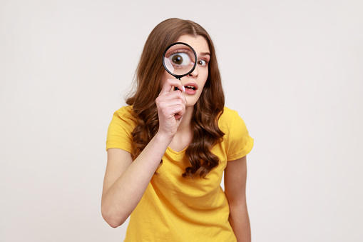 Portrait of funny young female in yellow casual T-shirt holding magnifying glass and looking at camera with big zoom eye, curious face. Indoor studio shot isolated on gray background.