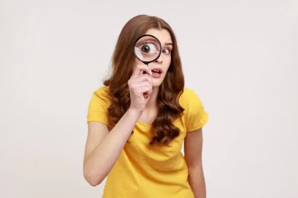 Photo of Portrait of funny young female in yellow casual T-shirt holding magnifying glass and looking at camera with big zoom eye, curious face.