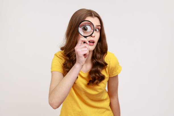 portrait of funny young female in yellow casual t-shirt holding magnifying glass and looking at camera with big zoom eye, curious face. - loupe quality control magnification glass imagens e fotografias de stock