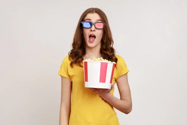 Astonished young female in 3d imax glasses watching movie film, hold popcorn, watching amazing movie, keeping mouth open, wears yellow T-shirt. Indoor studio shot isolated on gray background.