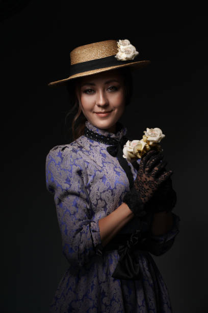 close-up de uma garota em pé em um vestido azul no estilo dos anos 1900, em um vestido retrô, chapéu e com um buquê de flores em um fundo cinza. moda histórica do século xix - women century 19th victorian style - fotografias e filmes do acervo