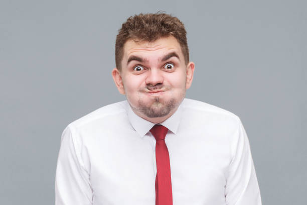 man blowing his cheeks and looking at camera with funny face and big eyes. funny portrait of young man in white shirt and tie standing blowing his cheeks and looking at camera with funny face and big eyes. indoor isolated on gray background. fool photos stock pictures, royalty-free photos & images