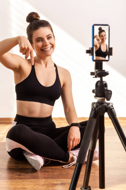 istock Online fitness trainer broadcasting workout online, points finger down, asking subscribe to her vlog 1346023602