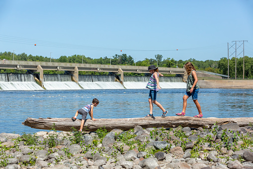 Three children (two sisters and their little brother) playing on the bank of the Mississippi River with the Coon Rapids, Minnesota, dam in the distance.