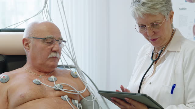 Cardiologist doctor using digital tablet talking with a senior patient on a ecg cycle in clinic