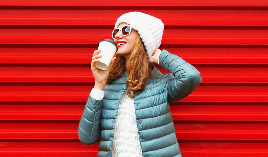 Portrait of happy caucasian smiling young woman with cup of coffee posing wearing a white hat on red background