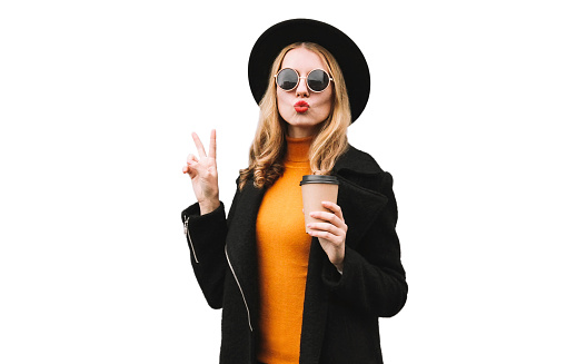 Portrait of stylish young woman with coffee cup blowing her lips wearing black coat and round hat isolated on white background