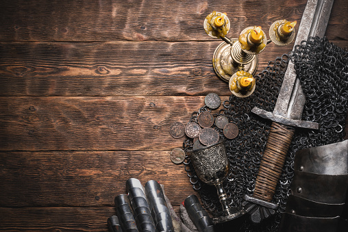 Knight sword and armor in the light of the burning candle on the wooden flat lay background with copy space.