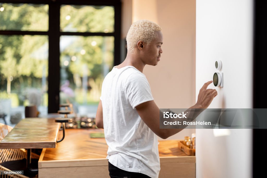 Latin man choosing temperature on thermostat Latin man choosing temperature on thermostat. Young focused guy pushing button on smart home system. Concept of modern domestic lifestyle. Interior of kitchen in modern apartment. Thermostat Stock Photo